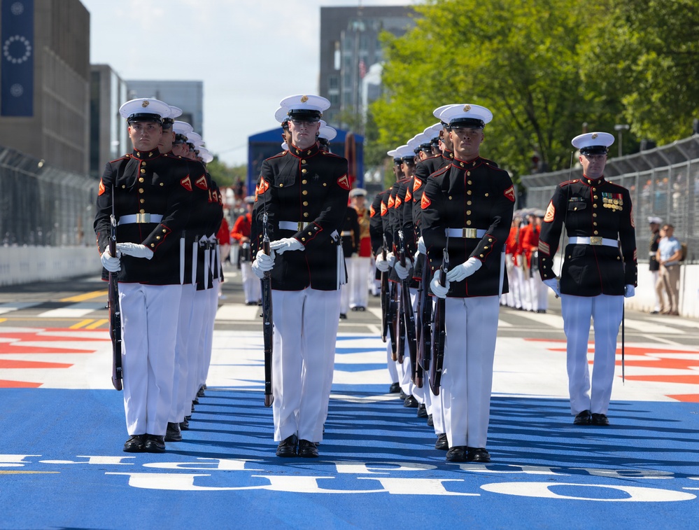 A formation of U.S. Marines in dress blues — black tunics with red trim, white trousers, and white covers — marches in precise formation along a blue-painted surface that appears to be a motorsport circuit or event venue. The Marines carry rifles and wear white gloves, indicating a ceremonial drill or honor guard performance. The setting appears to be an outdoor public event with spectators and barriers visible in the background.