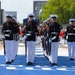 A formation of U.S. Marines in dress blues — black tunics with red trim, white trousers, and white covers — marches in precise formation along a blue-painted surface that appears to be a motorsport circuit or event venue. The Marines carry rifles and wear white gloves, indicating a ceremonial drill or honor guard performance. The setting appears to be an outdoor public event with spectators and barriers visible in the background.