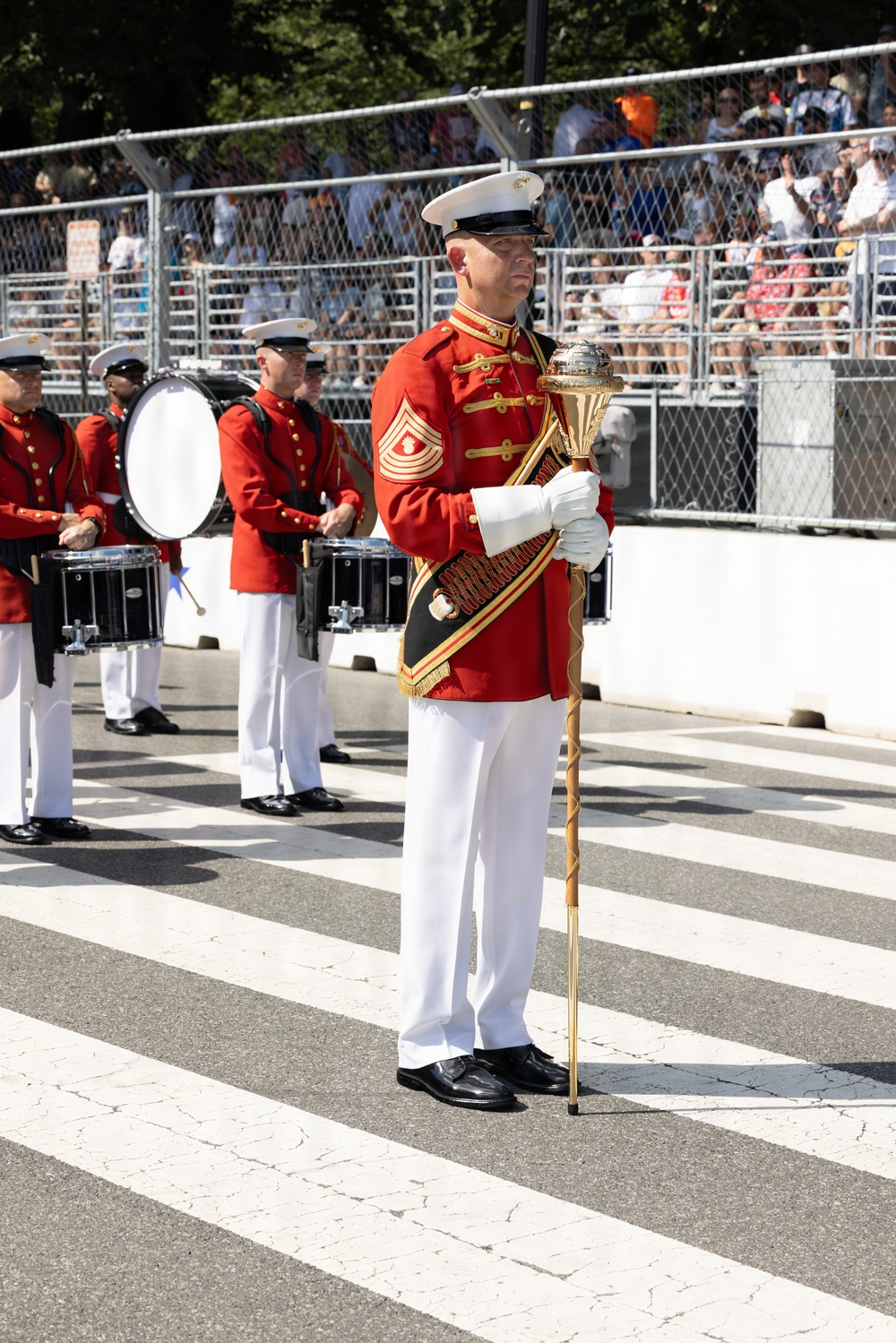 A United States Marine Corps band drum major stands at attention on a parade route, holding an ornate ceremonial mace with a gold and silver finial. He is dressed in the distinctive red full dress uniform with gold braiding, white trousers, white gloves, and a white peaked cap. Behind him, band members in matching red uniforms are positioned with snare drums and a bass drum, while spectators watch from bleachers in the background.