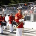 A United States Marine Corps band drum major stands at attention on a parade route, holding an ornate ceremonial mace with a gold and silver finial. He is dressed in the distinctive red full dress uniform with gold braiding, white trousers, white gloves, and a white peaked cap. Behind him, band members in matching red uniforms are positioned with snare drums and a bass drum, while spectators watch from bleachers in the background.