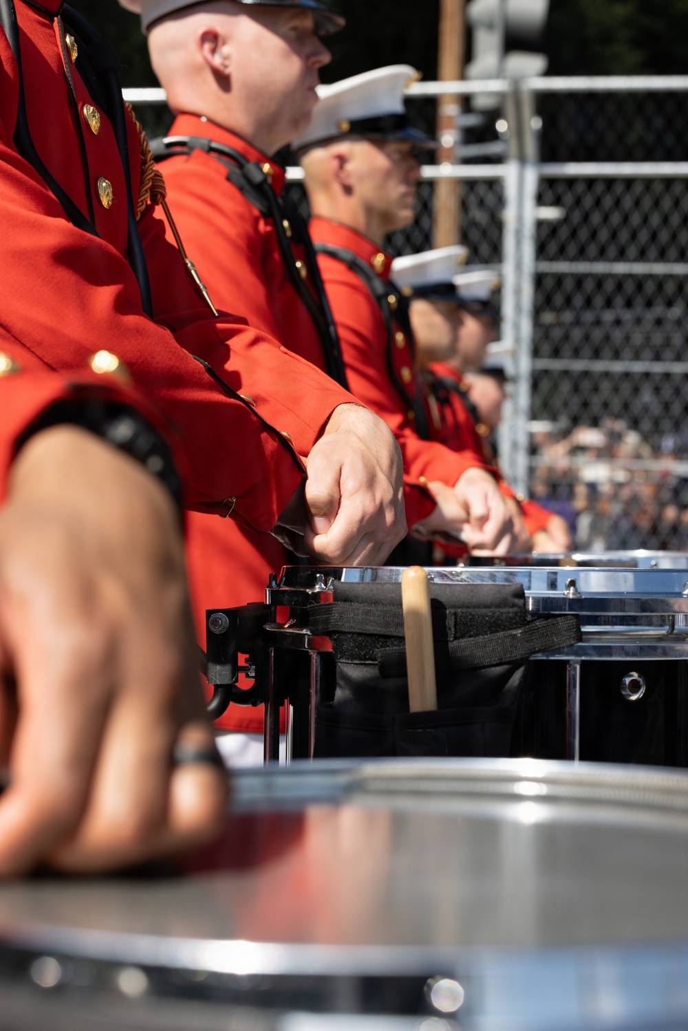 A line of military band drummers in red dress uniforms are performing at an outdoor event. The musicians are wearing scarlet tunics with gold buttons and black accents, consistent with U.S. Marine Corps ceremonial dress. Multiple snare drums with chrome hardware are visible in the foreground, with an audience and chain-link fence visible in the background.