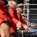 A line of military band drummers in red dress uniforms are performing at an outdoor event. The musicians are wearing scarlet tunics with gold buttons and black accents, consistent with U.S. Marine Corps ceremonial dress. Multiple snare drums with chrome hardware are visible in the foreground, with an audience and chain-link fence visible in the background.