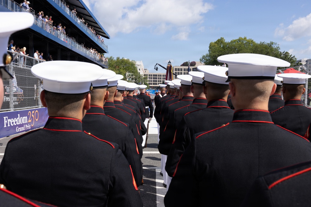 A formation of U.S. Marines in dress blues with white covers (caps) marches or stands at attention at an event branded as the 'Freedom 250 Grand Prix.' The scene takes place on a street circuit or parade route with multi-level spectator grandstands visible on the left side. The image is taken from behind the formation, showing the backs of the Marines' heads and uniforms.