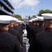 A formation of U.S. Marines in dress blues with white covers (caps) marches or stands at attention at an event branded as the 'Freedom 250 Grand Prix.' The scene takes place on a street circuit or parade route with multi-level spectator grandstands visible on the left side. The image is taken from behind the formation, showing the backs of the Marines' heads and uniforms.