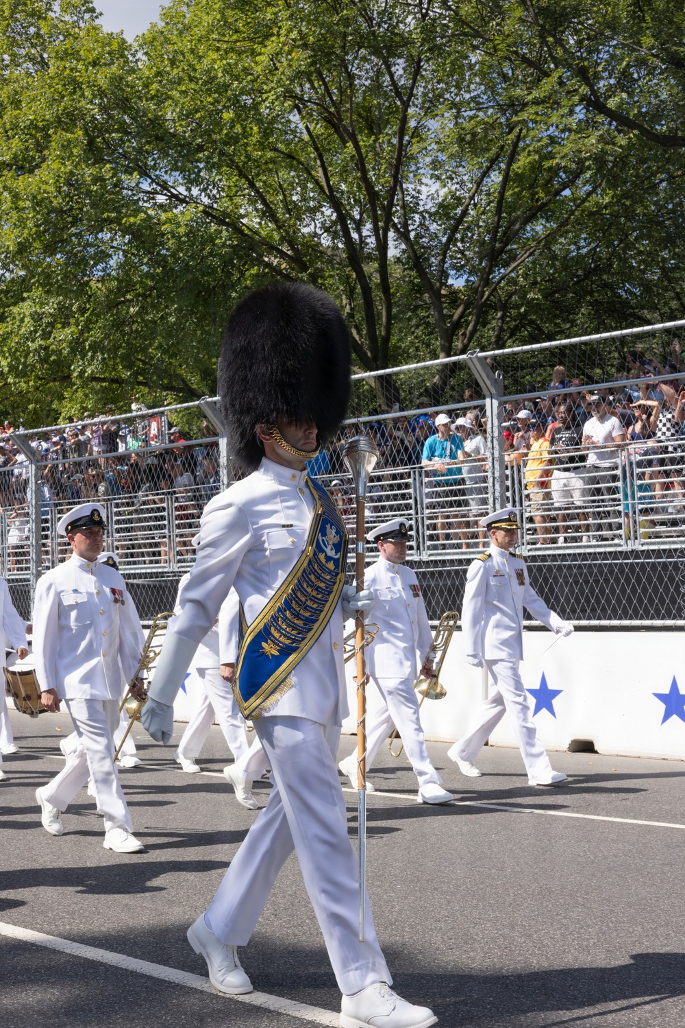A military marching band parade is taking place on a paved street in front of a crowd of spectators behind metal security fencing. The lead figure is a drum major wearing a distinctive tall black bearskin hat, white dress uniform, and a blue and gold ceremonial sash, carrying a mace. The setting appears to be an outdoor public event on a sunny day with lush green trees in the background.