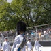 A military marching band parade is taking place on a paved street in front of a crowd of spectators behind metal security fencing. The lead figure is a drum major wearing a distinctive tall black bearskin hat, white dress uniform, and a blue and gold ceremonial sash, carrying a mace. The setting appears to be an outdoor public event on a sunny day with lush green trees in the background.