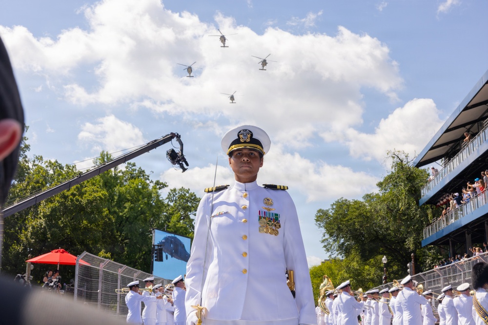 A U.S. Navy officer in dress whites stands at attention during a formal military ceremony or parade, with a military band visible behind her. Four military helicopters fly in formation overhead against a partly cloudy sky. The event appears to be a large public ceremony with spectators visible in bleachers to the right.