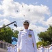 A U.S. Navy officer in dress whites stands at attention during a formal military ceremony or parade, with a military band visible behind her. Four military helicopters fly in formation overhead against a partly cloudy sky. The event appears to be a large public ceremony with spectators visible in bleachers to the right.