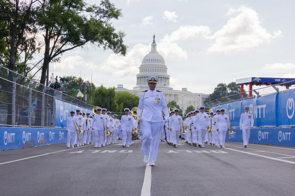 A military marching band in white dress uniforms parades down a closed street in Washington D.C., with the United States Capitol Building prominently visible in the background. The procession is led by a senior officer and takes place within a race circuit marked by blue NTT-branded barriers, suggesting a motorsport event pre-show or ceremonial opening. The scene combines military pageantry with a sporting event atmosphere.