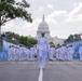 A military marching band in white dress uniforms parades down a closed street in Washington D.C., with the United States Capitol Building prominently visible in the background. The procession is led by a senior officer and takes place within a race circuit marked by blue NTT-branded barriers, suggesting a motorsport event pre-show or ceremonial opening. The scene combines military pageantry with a sporting event atmosphere.