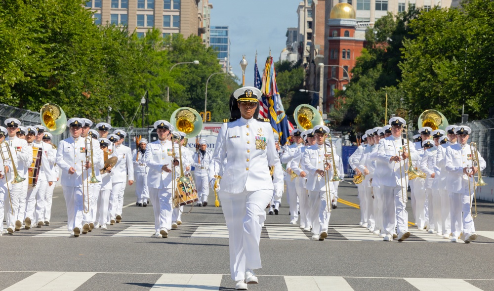 A military marching band parade is taking place on a city street, led by a female officer in a white dress uniform with gold insignia and medals. The band members, all dressed in white Navy-style uniforms, are playing brass instruments including trombones and sousaphones while marching in formation. Color guard members carrying American and military flags are visible in the background.