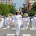 A military marching band parade is taking place on a city street, led by a female officer in a white dress uniform with gold insignia and medals. The band members, all dressed in white Navy-style uniforms, are playing brass instruments including trombones and sousaphones while marching in formation. Color guard members carrying American and military flags are visible in the background.