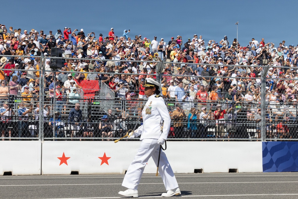 A military officer in white dress uniform marches along a track or parade route in front of a large, packed grandstand crowd. The officer is carrying a ceremonial sword or baton and wearing medals, suggesting a formal military ceremony or pre-event parade. The setting appears to be a motorsport venue based on the track surface, barrier walls with star decorations, and tiered spectator stands.