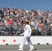 A military officer in white dress uniform marches along a track or parade route in front of a large, packed grandstand crowd. The officer is carrying a ceremonial sword or baton and wearing medals, suggesting a formal military ceremony or pre-event parade. The setting appears to be a motorsport venue based on the track surface, barrier walls with star decorations, and tiered spectator stands.