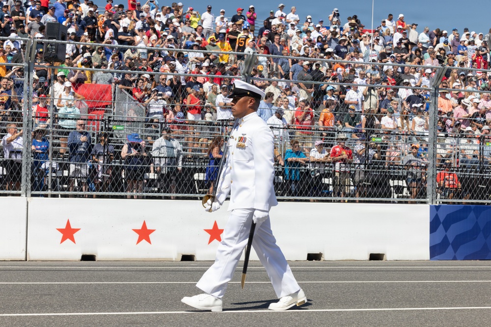 A U.S. Navy officer in full dress white uniform marches on a racetrack surface during a ceremonial event, carrying a ceremonial sword. The scene takes place at what appears to be a major motorsport venue, with a large packed grandstand crowd visible in the background. Red stars are visible on the pit lane wall, suggesting a patriotic or military ceremony at a racing event.