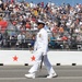 A U.S. Navy officer in full dress white uniform marches on a racetrack surface during a ceremonial event, carrying a ceremonial sword. The scene takes place at what appears to be a major motorsport venue, with a large packed grandstand crowd visible in the background. Red stars are visible on the pit lane wall, suggesting a patriotic or military ceremony at a racing event.