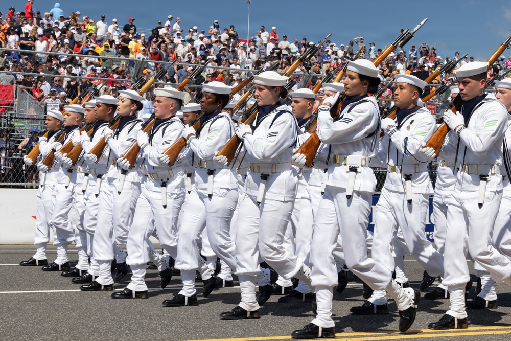 A precision drill team of U.S. Navy sailors in full dress white uniforms marches in formation during a public parade or ceremony. The sailors are carrying rifles at port arms in a synchronized manner, performing before a large crowd of spectators in stadium-style seating. The event appears to be a major public military demonstration held outdoors under clear skies.