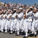 A precision drill team of U.S. Navy sailors in full dress white uniforms marches in formation during a public parade or ceremony. The sailors are carrying rifles at port arms in a synchronized manner, performing before a large crowd of spectators in stadium-style seating. The event appears to be a major public military demonstration held outdoors under clear skies.