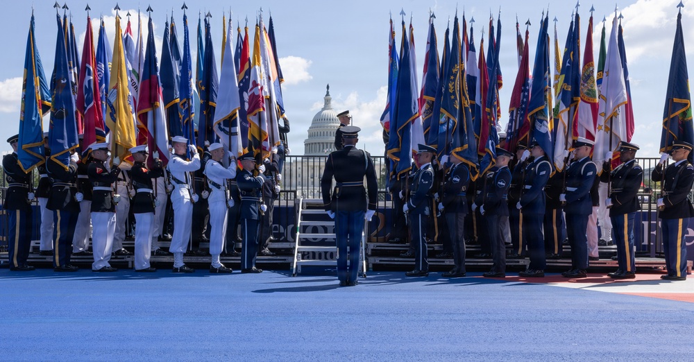 A formal military color guard ceremony is taking place on a blue-carpeted outdoor stage in front of the United States Capitol building in Washington, D.C. Multiple branches of the U.S. military are represented, with service members in distinct dress uniforms holding a large array of state and military flags. The event appears to be a high-profile national ceremony, likely an inauguration or major federal commemoration.