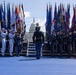 A formal military color guard ceremony is taking place on a blue-carpeted outdoor stage in front of the United States Capitol building in Washington, D.C. Multiple branches of the U.S. military are represented, with service members in distinct dress uniforms holding a large array of state and military flags. The event appears to be a high-profile national ceremony, likely an inauguration or major federal commemoration.