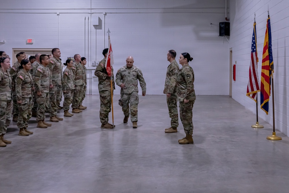 A military ceremony is taking place inside an indoor drill hall or armory, with soldiers in Army Combat Uniforms (ACU/OCP pattern) standing in formation. A central figure carrying a unit guidon (red flag) appears to be participating in a change of command or similar formal military ceremony. Three flags are displayed on the right side of the room, including the U.S. flag and what appears to be the Arizona state flag.