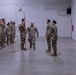 A military ceremony is taking place inside an indoor drill hall or armory, with soldiers in Army Combat Uniforms (ACU/OCP pattern) standing in formation. A central figure carrying a unit guidon (red flag) appears to be participating in a change of command or similar formal military ceremony. Three flags are displayed on the right side of the room, including the U.S. flag and what appears to be the Arizona state flag.