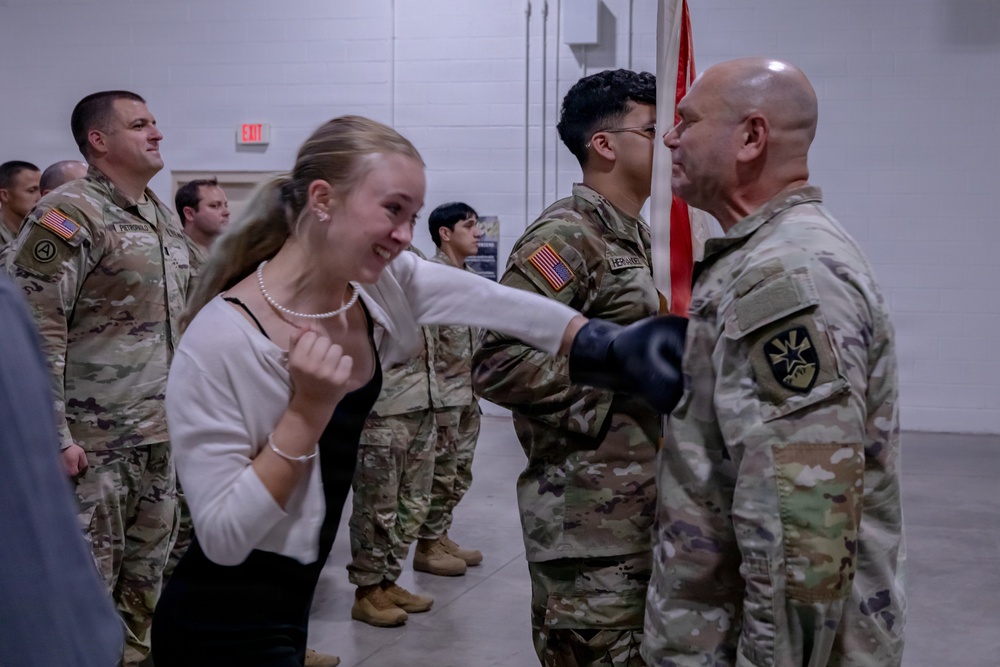 A military ceremony or event taking place indoors, likely a deployment or redeployment gathering. A young civilian woman in a white cardigan is smiling and reacting emotionally while a bald soldier in OCP (Operational Camouflage Pattern) uniform appears to be presenting or pinning something to her. Multiple soldiers in U.S. Army OCP uniforms are visible in the background.