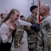 A military ceremony or event taking place indoors, likely a deployment or redeployment gathering. A young civilian woman in a white cardigan is smiling and reacting emotionally while a bald soldier in OCP (Operational Camouflage Pattern) uniform appears to be presenting or pinning something to her. Multiple soldiers in U.S. Army OCP uniforms are visible in the background.