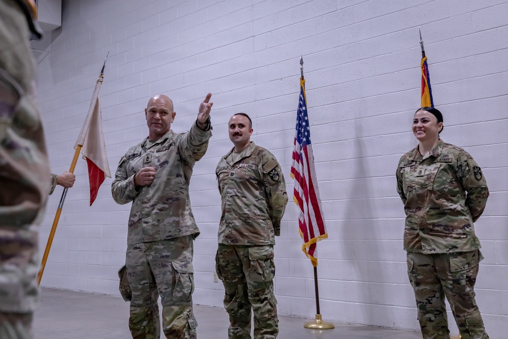 Three U.S. Army soldiers in MultiCam/OCP camouflage uniforms are standing in an indoor facility with a white cinder block wall background. The soldier on the left is holding a guidon (unit flag) and raising his right hand, suggesting an oath-taking or ceremonial event. Two flags are visible in the background: the U.S. national flag and what appears to be the Arizona state flag.