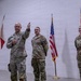 Three U.S. Army soldiers in MultiCam/OCP camouflage uniforms are standing in an indoor facility with a white cinder block wall background. The soldier on the left is holding a guidon (unit flag) and raising his right hand, suggesting an oath-taking or ceremonial event. Two flags are visible in the background: the U.S. national flag and what appears to be the Arizona state flag.