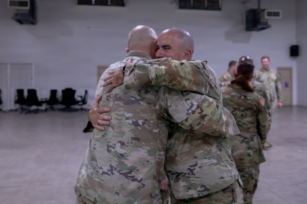 Two military personnel in camouflage uniforms are embracing in what appears to be a large indoor facility, likely a gymnasium or assembly hall. The emotional hug suggests a reunion or farewell scenario. Additional uniformed personnel are visible in the background engaged in similar interactions.