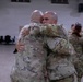 Two military personnel in camouflage uniforms are embracing in what appears to be a large indoor facility, likely a gymnasium or assembly hall. The emotional hug suggests a reunion or farewell scenario. Additional uniformed personnel are visible in the background engaged in similar interactions.