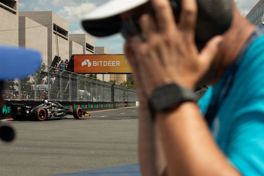 A photographer in a teal shirt and white cap captures a racing car on a street circuit track. In the background, a dark open-wheel race car with 'OpenAI' branding passes in front of a prominent orange 'BITDEER' sponsor banner. Spectators and safety fencing are visible along the track perimeter.
