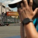 A photographer in a teal shirt and white cap captures a racing car on a street circuit track. In the background, a dark open-wheel race car with 'OpenAI' branding passes in front of a prominent orange 'BITDEER' sponsor banner. Spectators and safety fencing are visible along the track perimeter.