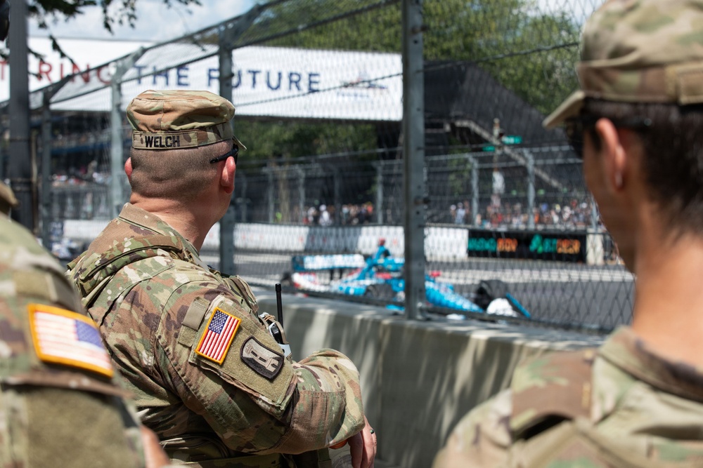 Two U.S. Army soldiers in OCP (Operational Camouflage Pattern) uniforms are watching what appears to be an IndyCar or Formula E street circuit race. The central soldier is identified by a name tape reading 'WELCH' and both wear American flag patches on their sleeves. They are positioned behind concrete barriers and chain-link fencing at the edge of the race track.