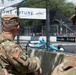Two U.S. Army soldiers in OCP (Operational Camouflage Pattern) uniforms are watching what appears to be an IndyCar or Formula E street circuit race. The central soldier is identified by a name tape reading 'WELCH' and both wear American flag patches on their sleeves. They are positioned behind concrete barriers and chain-link fencing at the edge of the race track.