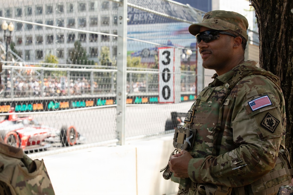 A U.S. Army soldier in MultiCam (OCP) camouflage uniform stands at what appears to be a street circuit motorsport racing event, likely providing security or ceremonial presence. The soldier wears an American flag patch and a unit patch on the right sleeve, sunglasses, and a patrol cap, and appears to be observing the race. A red racing car is visible on the track in the background, along with spectator stands and safety fencing.