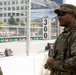 A U.S. Army soldier in MultiCam (OCP) camouflage uniform stands at what appears to be a street circuit motorsport racing event, likely providing security or ceremonial presence. The soldier wears an American flag patch and a unit patch on the right sleeve, sunglasses, and a patrol cap, and appears to be observing the race. A red racing car is visible on the track in the background, along with spectator stands and safety fencing.