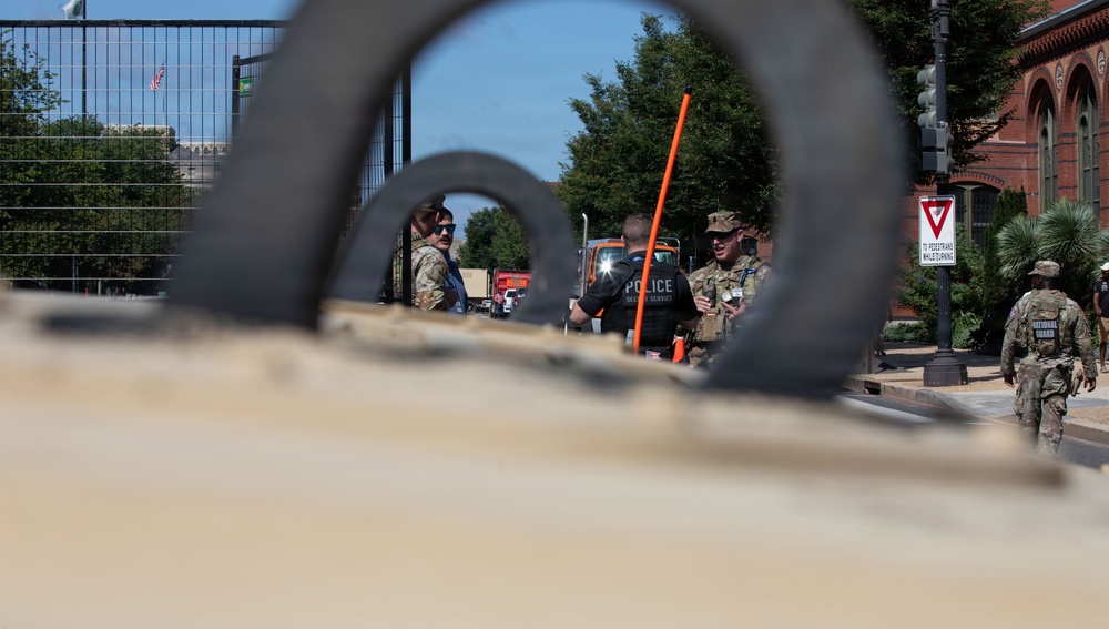 The image is taken through a curved metal barrier or gate structure, showing a security checkpoint scene. Multiple personnel are visible including Secret Service Police and National Guard soldiers in military camouflage uniforms. The setting appears to be a secured perimeter near a government or institutional area, identifiable by the red brick building and fencing in the background.