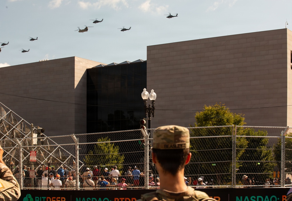 A military flyover featuring multiple helicopters, including what appear to be CH-47 Chinooks and AH-64 Apaches, passes over an urban area during a public event. A soldier with the name tape 'BEASLEE' visible on their uniform observes the flyover from behind a security fence. A large crowd of spectators is gathered behind barriers, with a modern stone building and NASDAQ-branded signage visible in the background.
