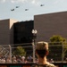 A military flyover featuring multiple helicopters, including what appear to be CH-47 Chinooks and AH-64 Apaches, passes over an urban area during a public event. A soldier with the name tape 'BEASLEE' visible on their uniform observes the flyover from behind a security fence. A large crowd of spectators is gathered behind barriers, with a modern stone building and NASDAQ-branded signage visible in the background.