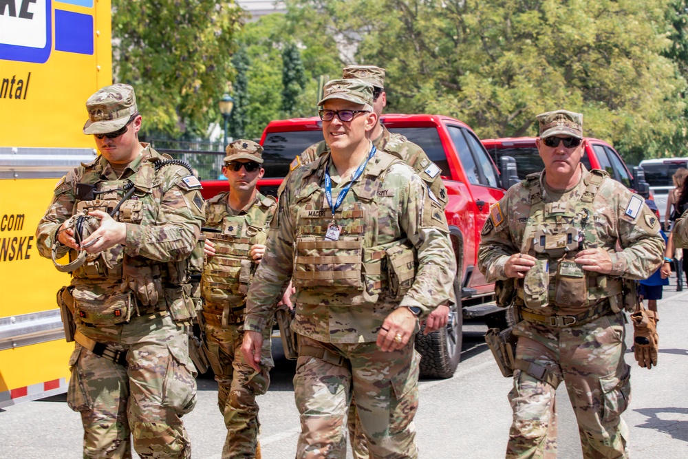 A group of U.S. Army soldiers in OCP (Operational Camouflage Pattern) uniforms are walking together in what appears to be an outdoor urban setting. The central figure is wearing glasses, a blue lanyard with an ID badge, and a tactical vest, suggesting a senior officer or official role. The setting appears to be a public event or security detail based on the civilian vehicles and surroundings.