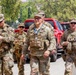 A group of U.S. Army soldiers in OCP (Operational Camouflage Pattern) uniforms are walking together in what appears to be an outdoor urban setting. The central figure is wearing glasses, a blue lanyard with an ID badge, and a tactical vest, suggesting a senior officer or official role. The setting appears to be a public event or security detail based on the civilian vehicles and surroundings.