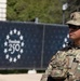 A U.S. Army soldier in full combat uniform and tactical gear stands at attention, appearing to be on security duty. The soldier, identified by name tape as 'PITTMAN,' is positioned in front of a dark barrier displaying a 'Freedom 250' logo surrounded by a circle of stars. The setting suggests a security or ceremonial post at a formal event or installation.