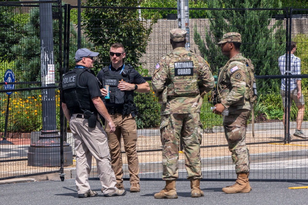 Four security personnel are gathered near a black metal security fence in an outdoor setting. Two are identified as U.S. Secret Service agents wearing tactical vests labeled 'Secret Service' and 'Police Secret Service,' while two others are U.S. National Guard soldiers in camouflage uniforms. The group appears to be engaged in a briefing or coordination discussion.