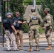 Four security personnel are gathered near a black metal security fence in an outdoor setting. Two are identified as U.S. Secret Service agents wearing tactical vests labeled 'Secret Service' and 'Police Secret Service,' while two others are U.S. National Guard soldiers in camouflage uniforms. The group appears to be engaged in a briefing or coordination discussion.