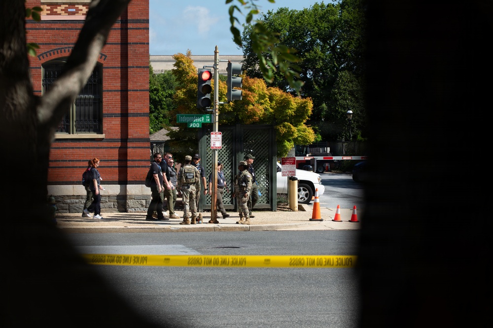The image shows a law enforcement response scene on Independence Avenue, viewed through tree branches in the foreground. Yellow police tape reading 'POLICE LINE DO NOT CROSS' is stretched across the street, with multiple armed personnel in tactical gear gathered on the sidewalk. Orange traffic cones and a security barrier are visible near what appears to be a secured government facility entrance.