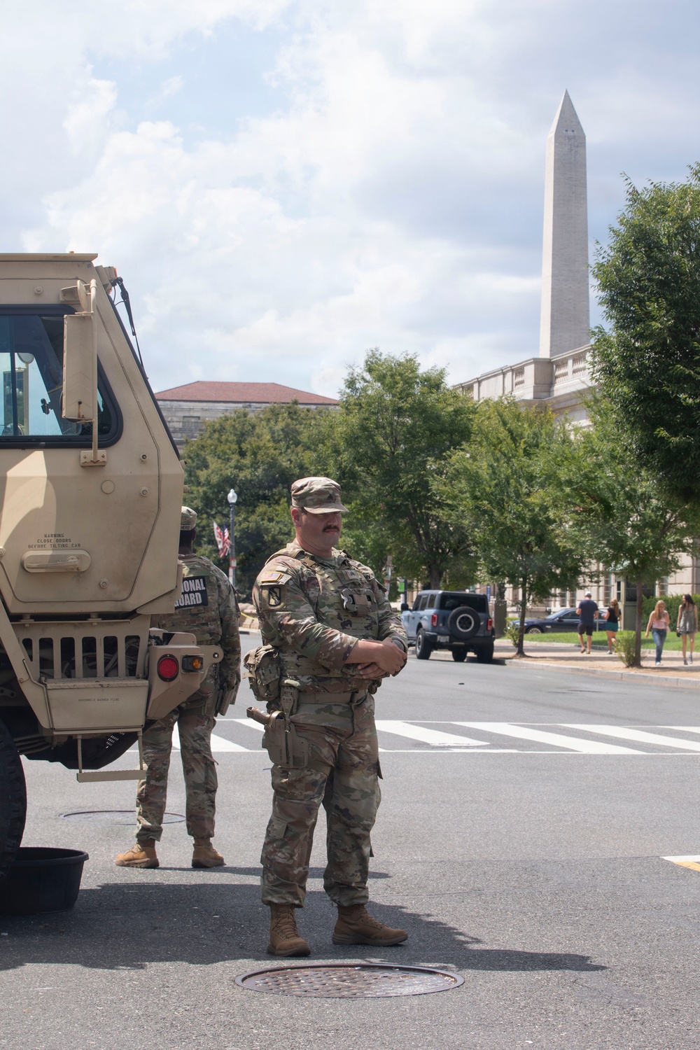 Two U.S. National Guard soldiers in OCP (Operational Camouflage Pattern) uniforms stand beside a tan military tactical vehicle on a street in Washington, D.C. The Washington Monument is clearly visible in the background, along with neoclassical government buildings and civilian pedestrians. The scene suggests a security deployment or civil support mission in the nation's capital.