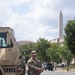Two U.S. National Guard soldiers in OCP (Operational Camouflage Pattern) uniforms stand beside a tan military tactical vehicle on a street in Washington, D.C. The Washington Monument is clearly visible in the background, along with neoclassical government buildings and civilian pedestrians. The scene suggests a security deployment or civil support mission in the nation's capital.