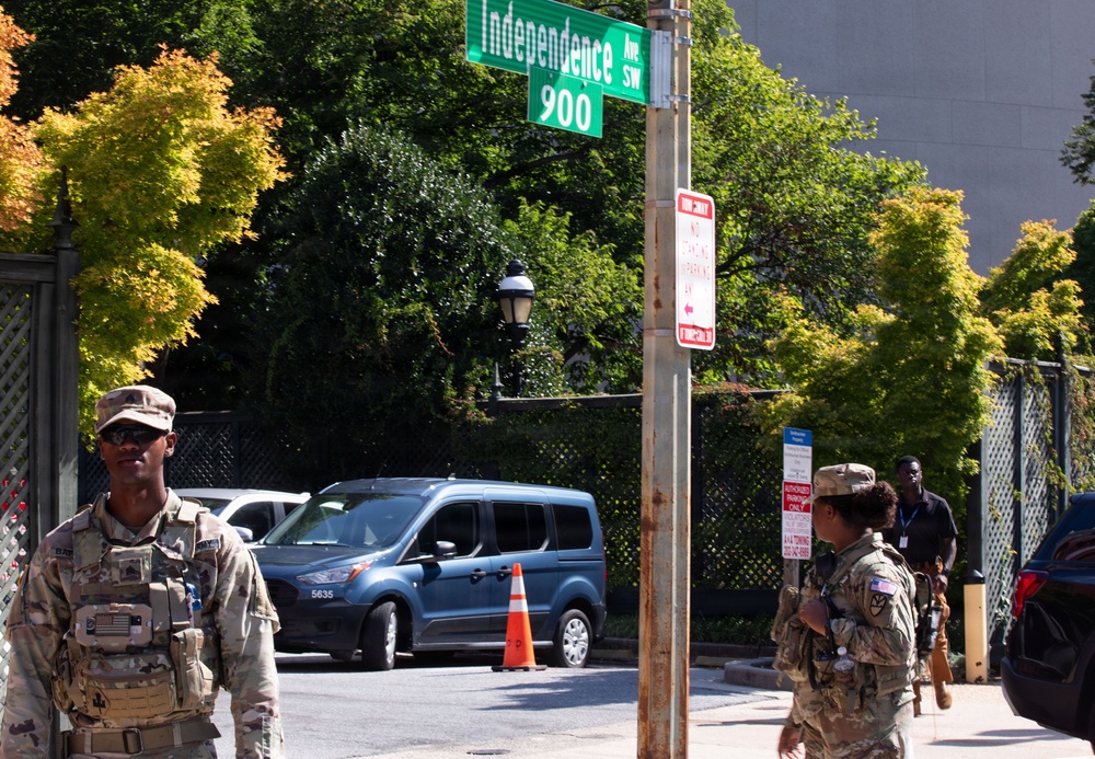 Two armed military personnel in camouflage uniforms are stationed at the intersection of Independence Avenue SW at the 900 block, likely in Washington D.C. They appear to be providing security at what looks like a controlled access point, with a traffic cone blocking vehicle entry. A civilian with a badge/lanyard is visible in the background near a building entrance.